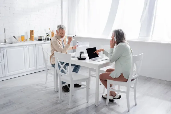 Hombre sosteniendo facturas y señalando a la esposa hablando en el teléfono inteligente cerca de computadoras portátiles en casa - foto de stock