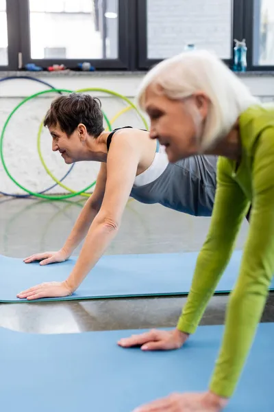 Vista lateral de mulheres idosas sorridentes fazendo prancha em tapetes de fitness no ginásio — Fotografia de Stock