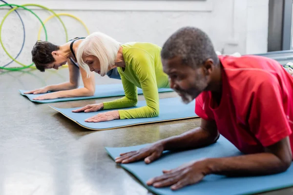 Mulher sênior exercitando no tapete de fitness perto de pessoas inter-raciais fazendo prancha no ginásio — Fotografia de Stock