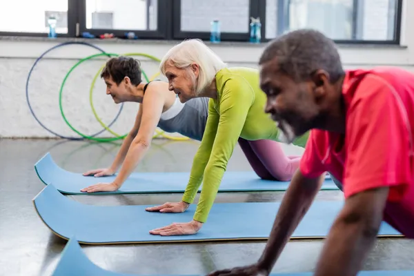 Side view of smiling elderly woman exercising near multiethnic people in sports center — Stock Photo