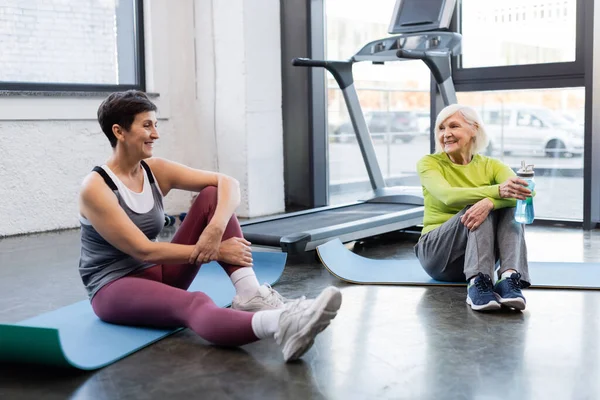 Mujeres mayores sonrientes con botella de deporte descansando en colchonetas de fitness en el centro deportivo — Stock Photo