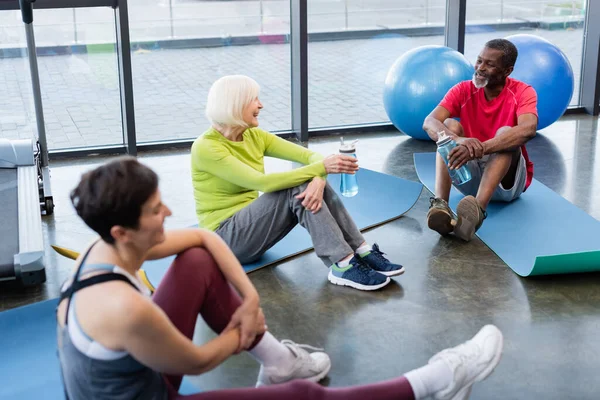 Cheerful senior interracial people sitting on fitness mats in sports center — Stock Photo