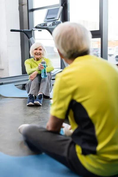 Femme âgée souriante tenant une bouteille de sport près de l'homme flou dans la salle de gym — Photo de stock