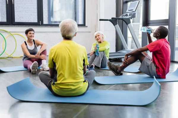 Positive multiethnic senior people with sports bottles resting on fitness mats in gym — Stock Photo