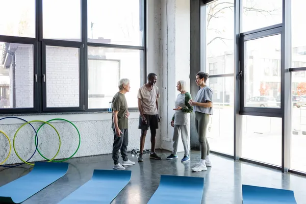 Interrassische Senioren in Sportkleidung unterhalten sich im Fitnessstudio — Stockfoto