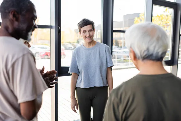 Femme âgée souriante debout près des amis interracial dans la salle de gym — Photo de stock