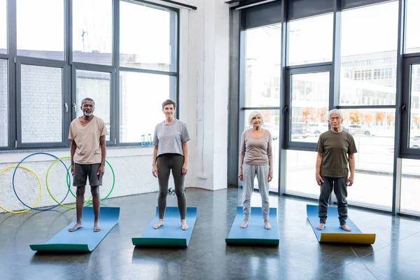 Personas mayores interraciales en ropa deportiva de pie sobre alfombras de fitness en el gimnasio - foto de stock