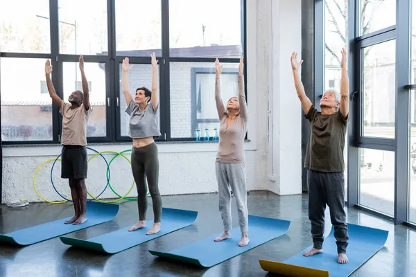 Personas mayores multiétnicas levantando las manos durante la práctica de yoga en el gimnasio - foto de stock