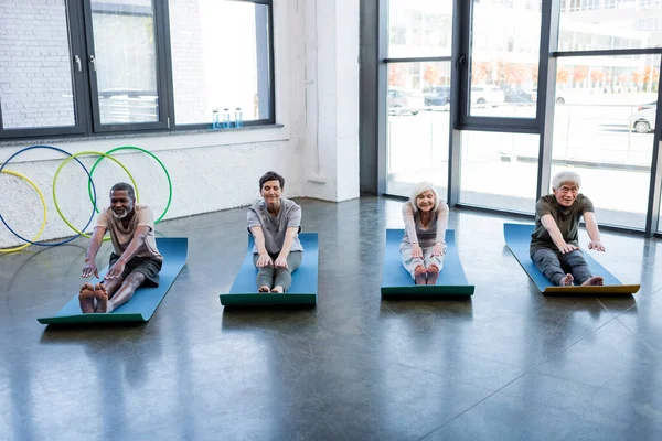 Positivo interrazziale anziani stretching su tappetini fitness in palestra — Foto stock