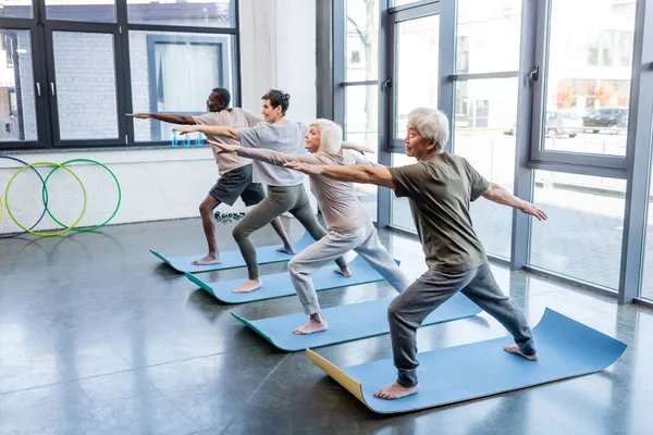 Hombre asiático senior en pose guerrera practicando yoga cerca de amigos multiétnicos en centro deportivo - foto de stock