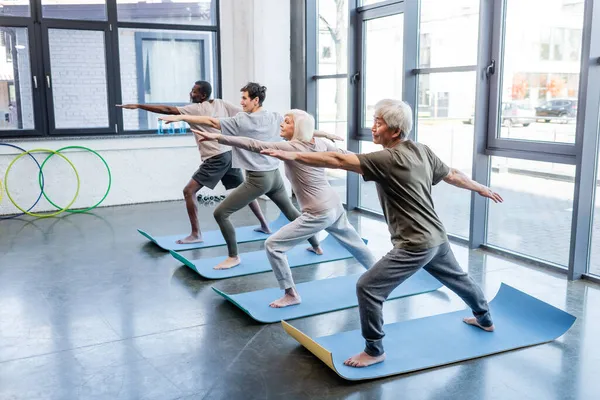 Senior asian man in warrior pose practicing yoga near multiethnic friends in sports center — Stock Photo