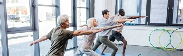 Interracial senior people doing warrior pose in sports center, banner — Stock Photo