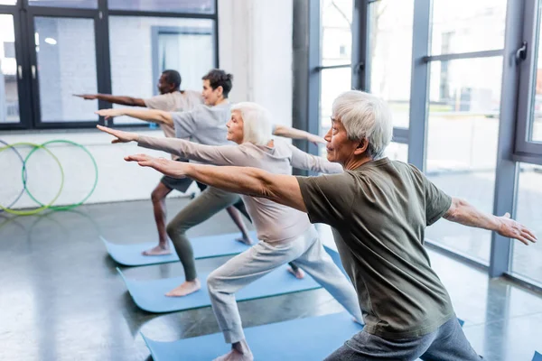 Anciano asiático practicando yoga cerca de personas multiétnicas en centro deportivo - foto de stock