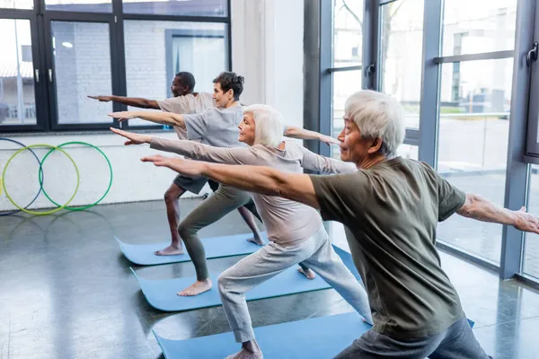 Mujer mayor en pose guerrera practicando yoga cerca de personas multiétnicas en el gimnasio - foto de stock