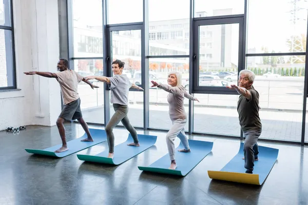 Multicultural elderly people standing in warrior pose on mats in sports center — Stock Photo