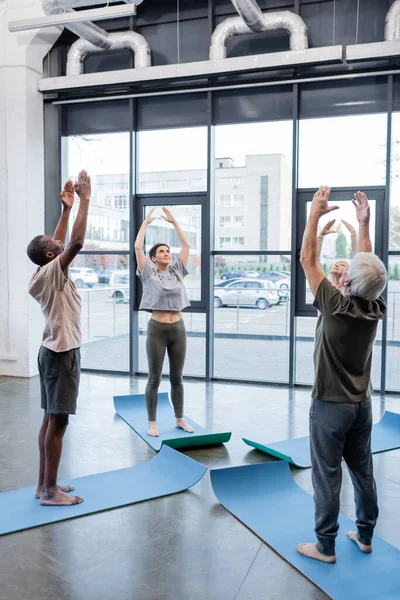 Personas mayores interraciales levantando las manos mientras practican yoga en esteras en un centro deportivo - foto de stock