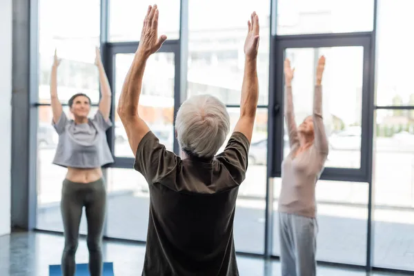 Senior man raising hands while practicing yoga near blurred friends in sports center — Stock Photo