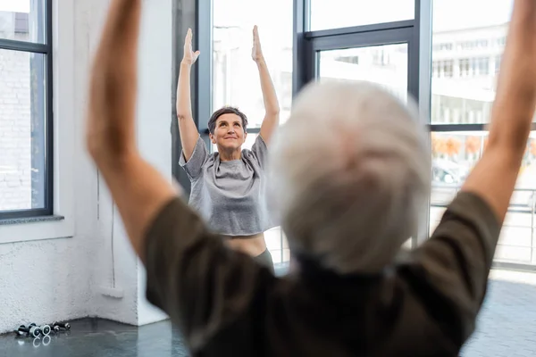 Mujer mayor sonriente levantando las manos durante la práctica de yoga cerca del hombre borroso en el centro deportivo - foto de stock