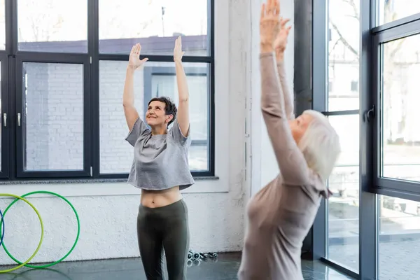 Happy senior woman practicing mountain pose near blurred friend in sports center — Stock Photo