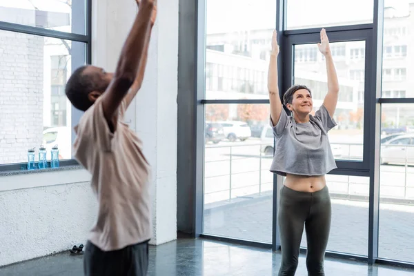 Fröhliche Seniorin posiert in Bergpose in der Nähe eines Afroamerikaners im Sportzentrum — Stockfoto