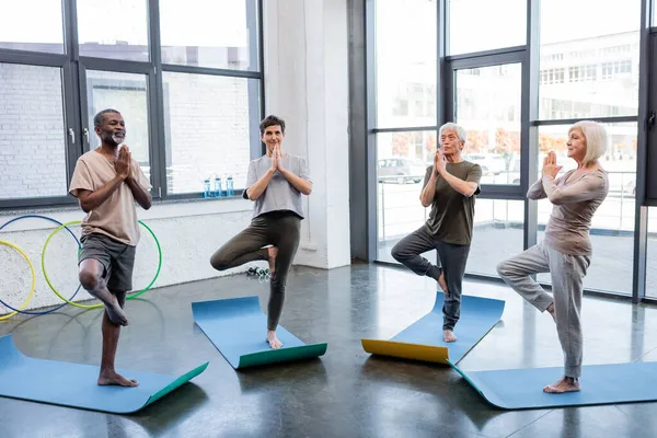 Interracial senior people standing in tree pose on yoga mats in sports center — Stock Photo