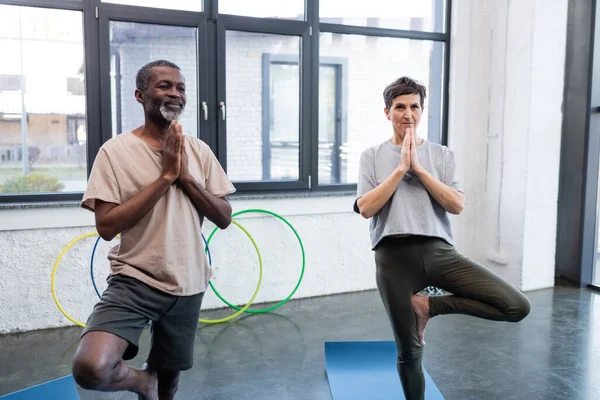 Elderly interracial people standing in yoga tree pose in sports center — Stock Photo