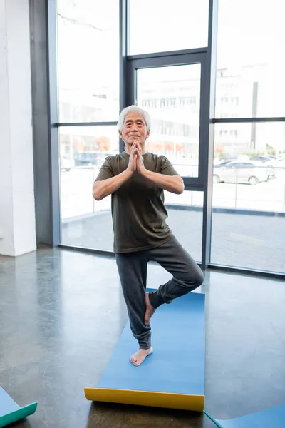 Anciano asiático hombre de pie en árbol pose en yoga mat en centro deportivo - foto de stock