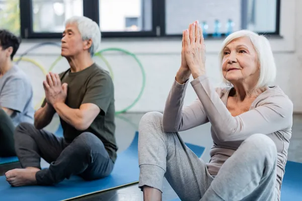 Mujer de pelo gris practicando yoga cerca borrosa asiática amiga en centro deportivo - foto de stock