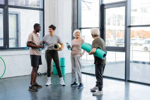 Heureuses personnes âgées multiethniques avec des bouteilles de sport et des tapis de fitness parlant dans la salle de gym — Photo de stock