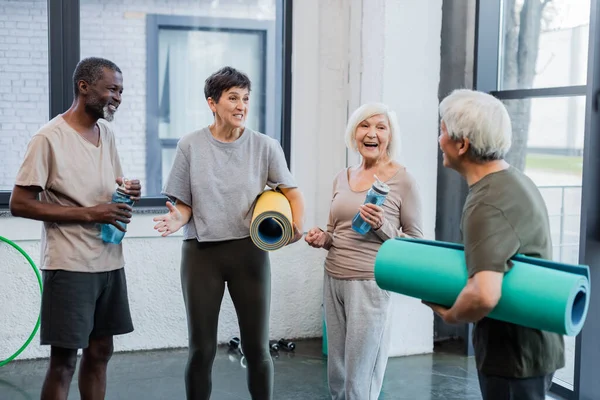 Positive Sportlerin mit Fitnessmatte im Gespräch mit multiethnischen Freunden im Fitnessstudio — Stockfoto