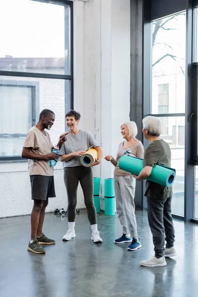 Homme afro-américain avec bouteille de sport pointant vers des amis multiethniques avec des tapis de fitness dans le centre sportif — Photo de stock