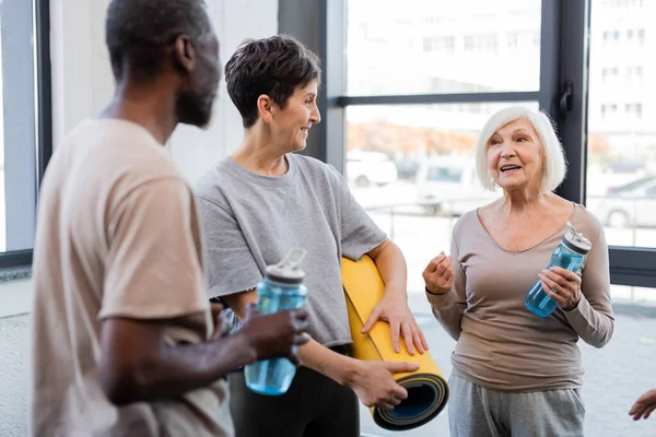 Femme âgée tenant bouteille de sport et parlant à des amis interraciaux dans la salle de gym — Photo de stock