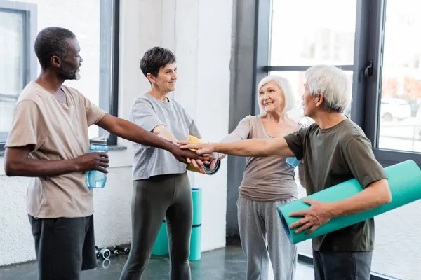 Personas mayores interracial sosteniendo alfombras de fitness y manos en el gimnasio - foto de stock