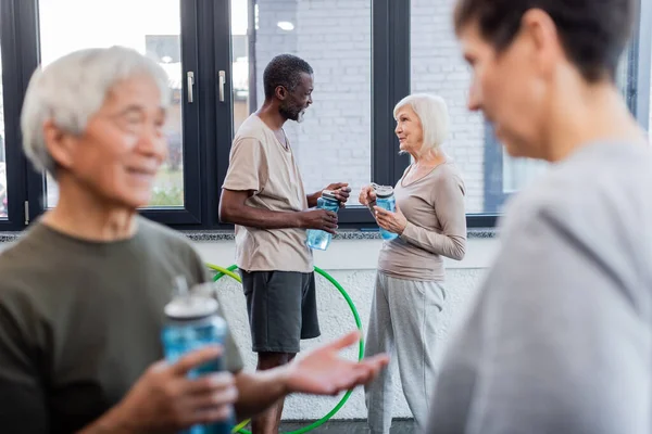 Personnes âgées multiethniques tenant des bouteilles de sport tout en parlant dans la salle de gym — Photo de stock