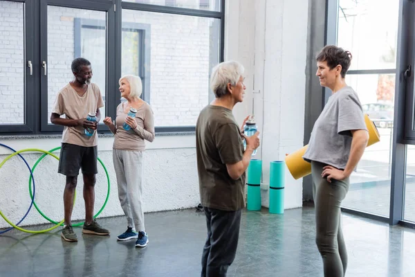 Positiv multikulturelle Menschen in Sportbekleidung, die Sportflaschen in der Hand halten, während sie im Fitnessstudio reden — Stockfoto