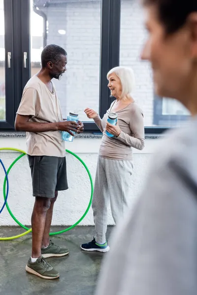 Gente multiétnica sonriente con botellas deportivas hablando en el gimnasio - foto de stock