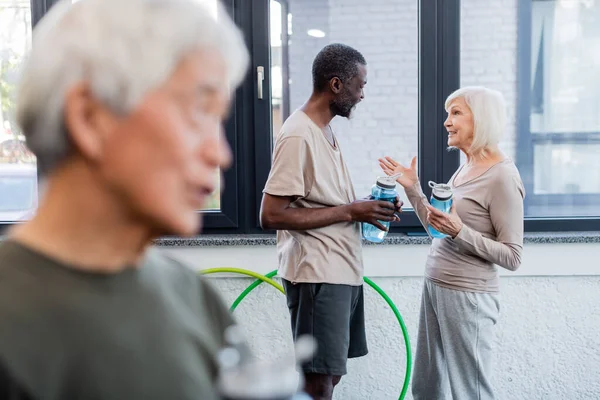 Gray haired woman with sports bottle talking to african american man in sports center — Stock Photo