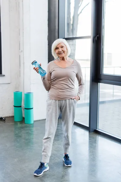 Deportiva de edad avanzada positiva sosteniendo botella deportiva en el gimnasio - foto de stock