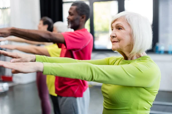 Elderly sportswoman training near blurred multiethnic people in sports center — Stock Photo