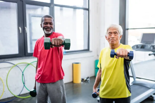 Hanteln in den Händen verschwommener multiethnischer Sportler, die im Fitnessstudio trainieren — Stockfoto