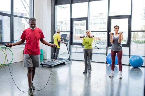 Entraînement sportif afro-américain avec corde à sauter près des sportives avec haltères dans la salle de gym — Photo de stock