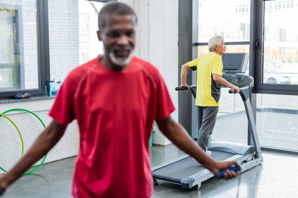 Senior asian man training on treadmill near blurred african american man with jump rope in gym — Stock Photo