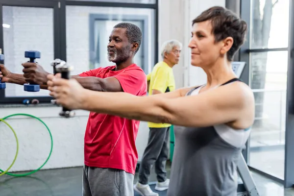 Deportista afroamericana haciendo ejercicio con pesas cerca de deportista senior en centro deportivo - foto de stock