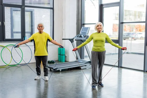 Senior mujer entrenamiento con saltar cuerda cerca asiático amigo en deportivo centro - foto de stock