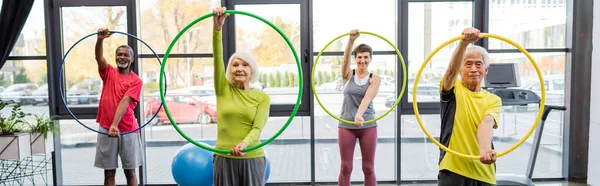 Grupo de pessoas idosas multiétnicas treinando com aros hula no centro esportivo, banner — Fotografia de Stock