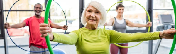 Sonriente mujer de pelo gris sosteniendo hula hoop mientras entrena en el gimnasio, pancarta - foto de stock