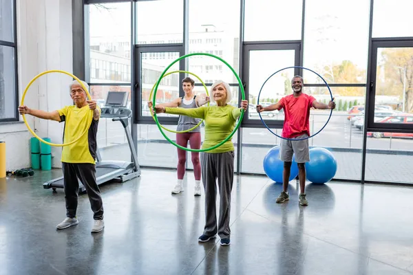 Pessoas idosas multiétnicas treinando com aros de hula perto de esteira e bolas de fitness no ginásio — Fotografia de Stock