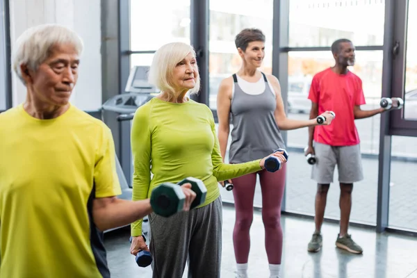 Lächelnde grauhaarige Frau trainiert mit Hanteln bei multikulturellen Freunden im Fitnessstudio — Stockfoto