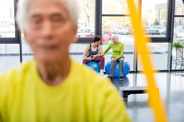 Smiling elderly sportswomen talking on fitness balls near blurred man training in gym — Stock Photo