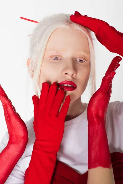 Bonito modelo albino en guantes rojos posando cerca de manos femeninas en pintura aislada sobre blanco - foto de stock